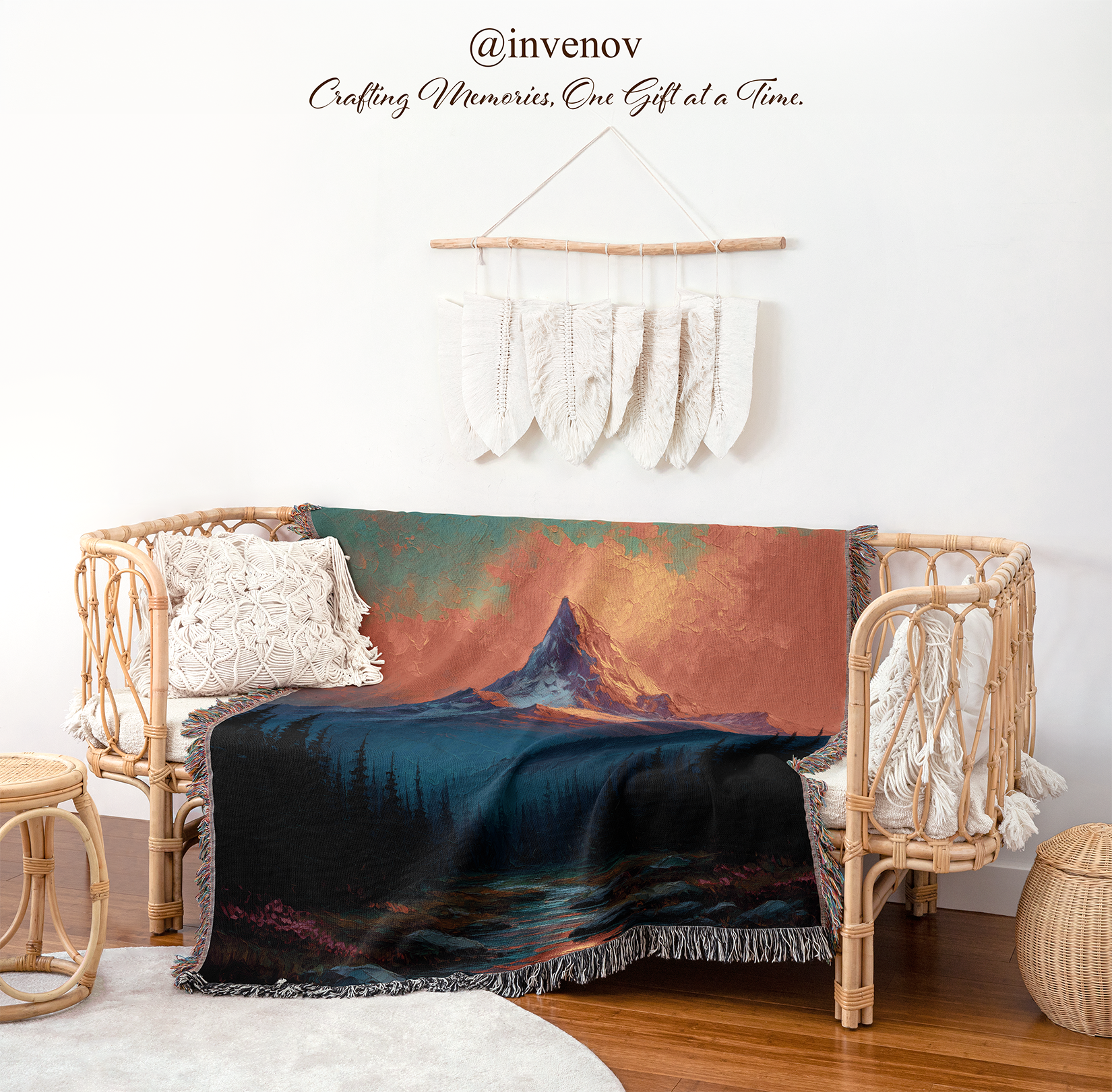 A woven throw blanket with a mountain landscape print is draped over a rattan daybed in a styled room. The blanket features a dark blue lake and forest under a warm orange and teal sky with a mountain peak.