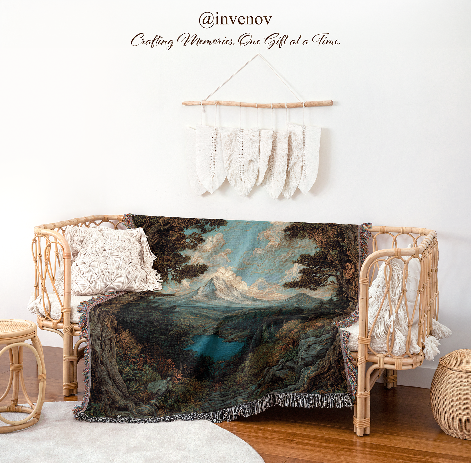 A woven landscape throw blanket with a mountain and lake scene is draped over a rattan daybed in a styled room. The scene includes a white wall, decorative macrame wall hanging, pillows, a small woven stool, and a round basket on a wood floor.