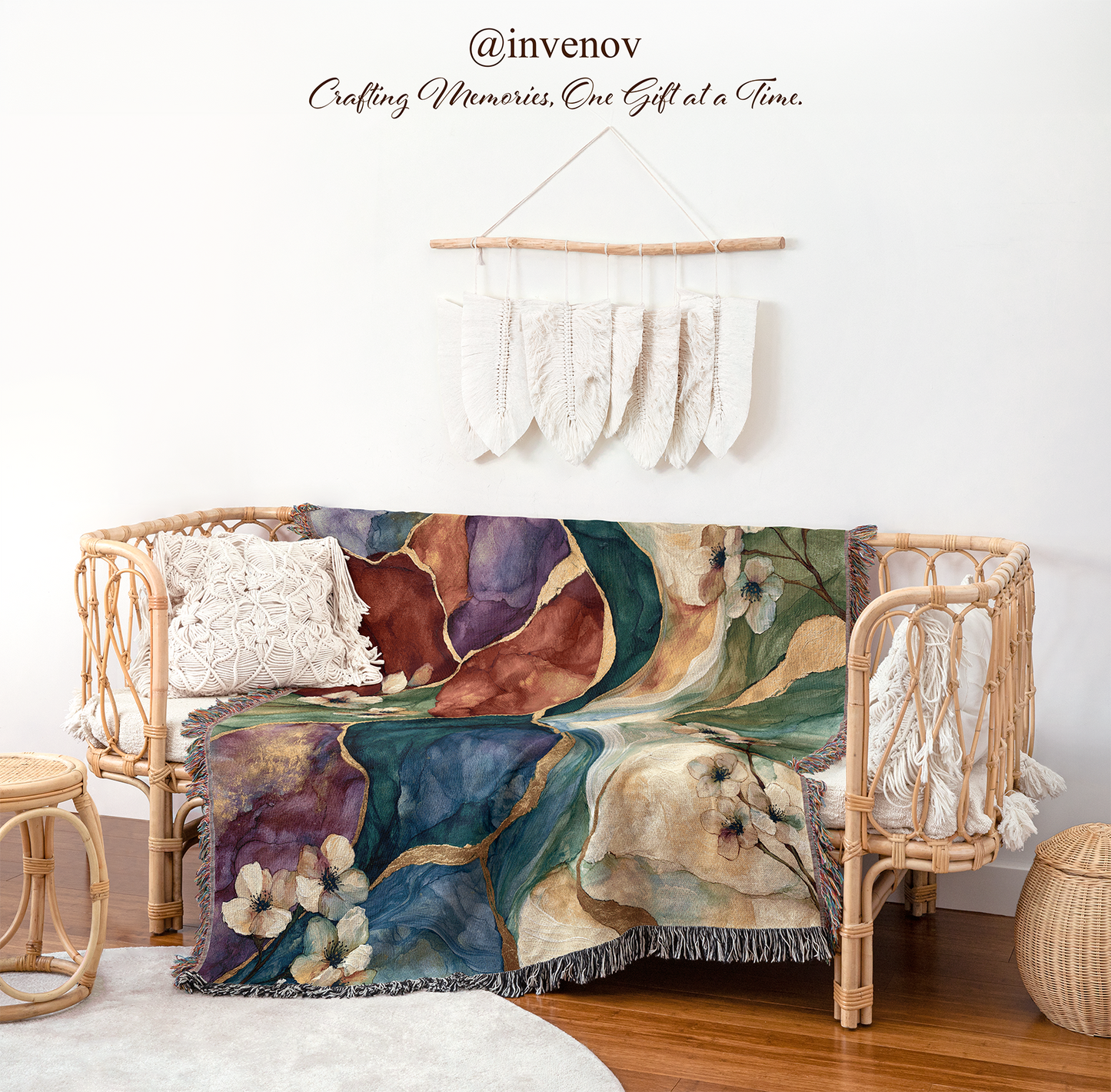 A woven blanket styled in a boho room setting, draped with a large abstract floral and landscape throw blanket. The blanket features a rich palette of deep teal, purple, brown, and cream with gold accents and a fringed edge.