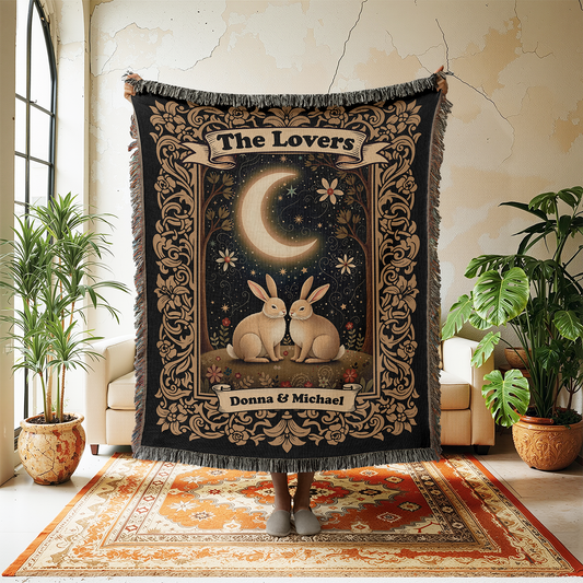 Decorative throw blanket with 'The Lovers' design featuring two rabbits and a moon, held up in a room with plants and furniture.