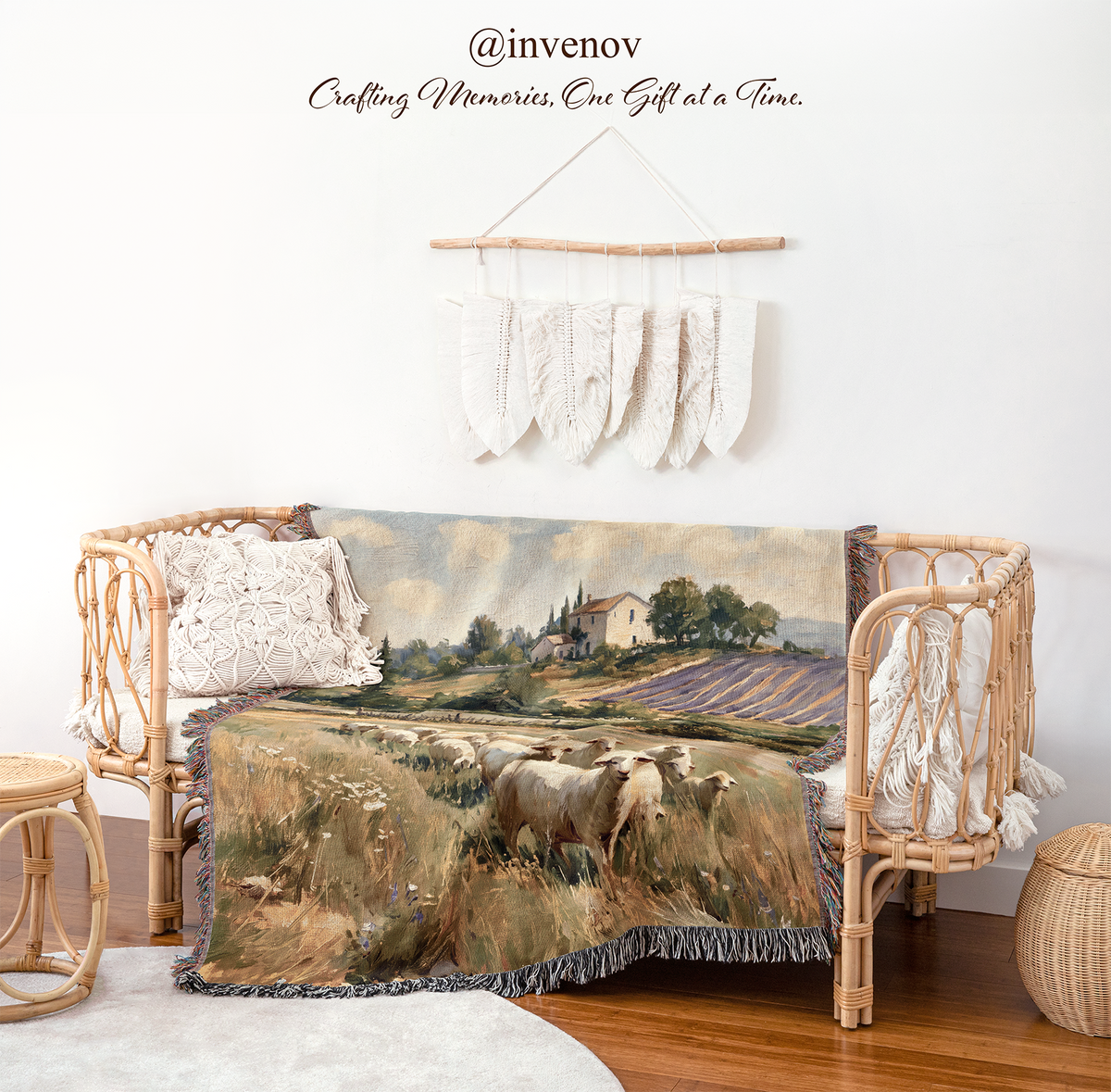 A woven rattan daybed styled in a bright, modern boho room, draped with a scenic landscape throw blanket featuring sheep in a field. The room has a white wall with a macrame wall hanging and the brand name '@invenov' printed on it, along with a small wooden stool and a round woven basket.