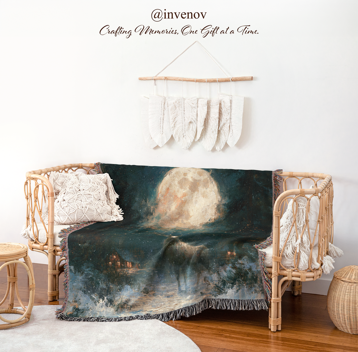 A dark navy and black blanket featuring a moonlit seascape with a large glowing moon, stars, and small house lights, draped over a rattan daybed in a styled room. The scene includes white woven pillows, a wall hanging, a small wooden stool, and a round woven side table.