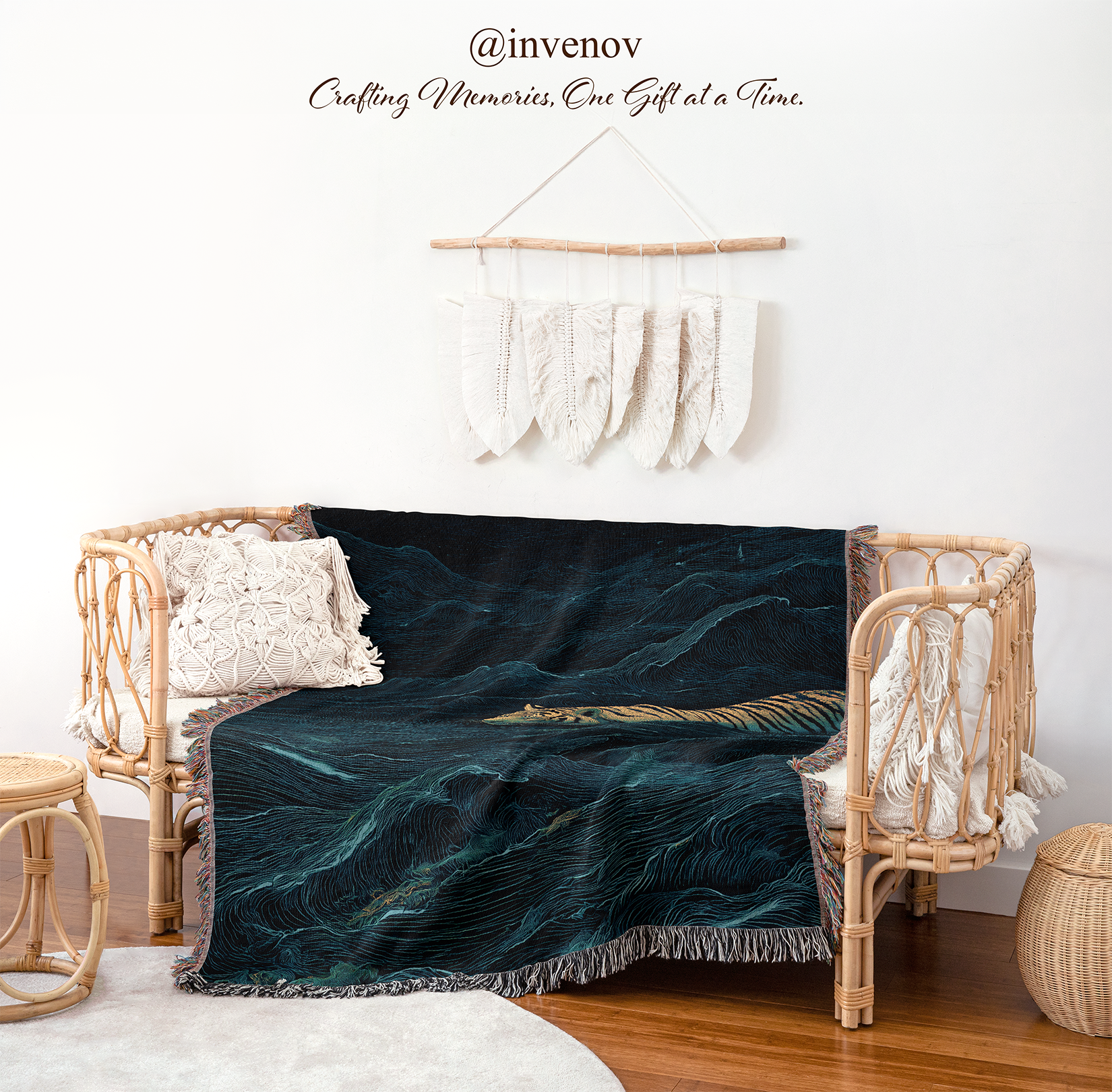 A woven rattan daybed in a styled room setting, draped with a dark teal and black mountain landscape throw blanket. The room features a white wall with a macrame wall hanging, a wooden floor, and a small woven stool.