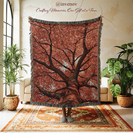A large woven throw blanket featuring a detailed tree design in earthy brown and rust tones, held up by a person standing on a patterned rug in a styled living room with plants and beige chairs.