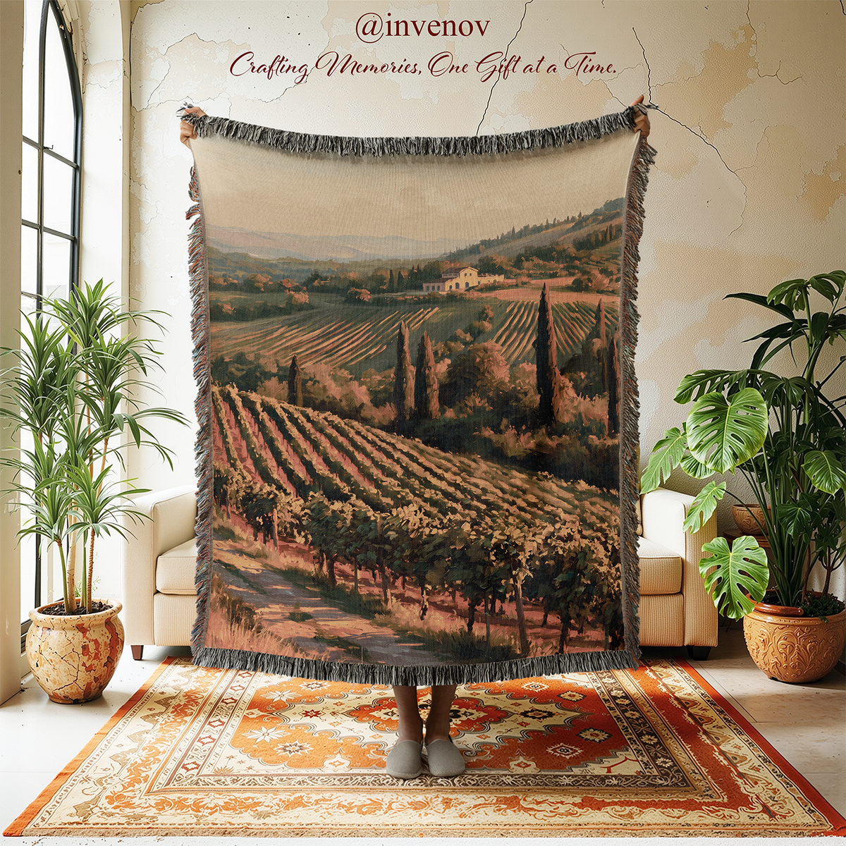 A large woven throw blanket with fringe edges is held up in a styled room. The blanket features a scenic Tuscan vineyard landscape print.