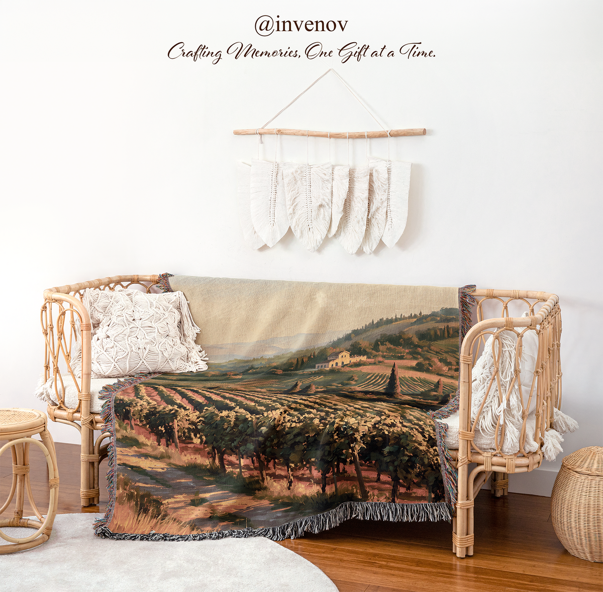 A woven vineyard landscape throw blanket is draped over a rattan daybed in a styled room setting. The blanket features a detailed print of rolling hills, trees, and grapevines in earthy tones of green, brown, and beige.