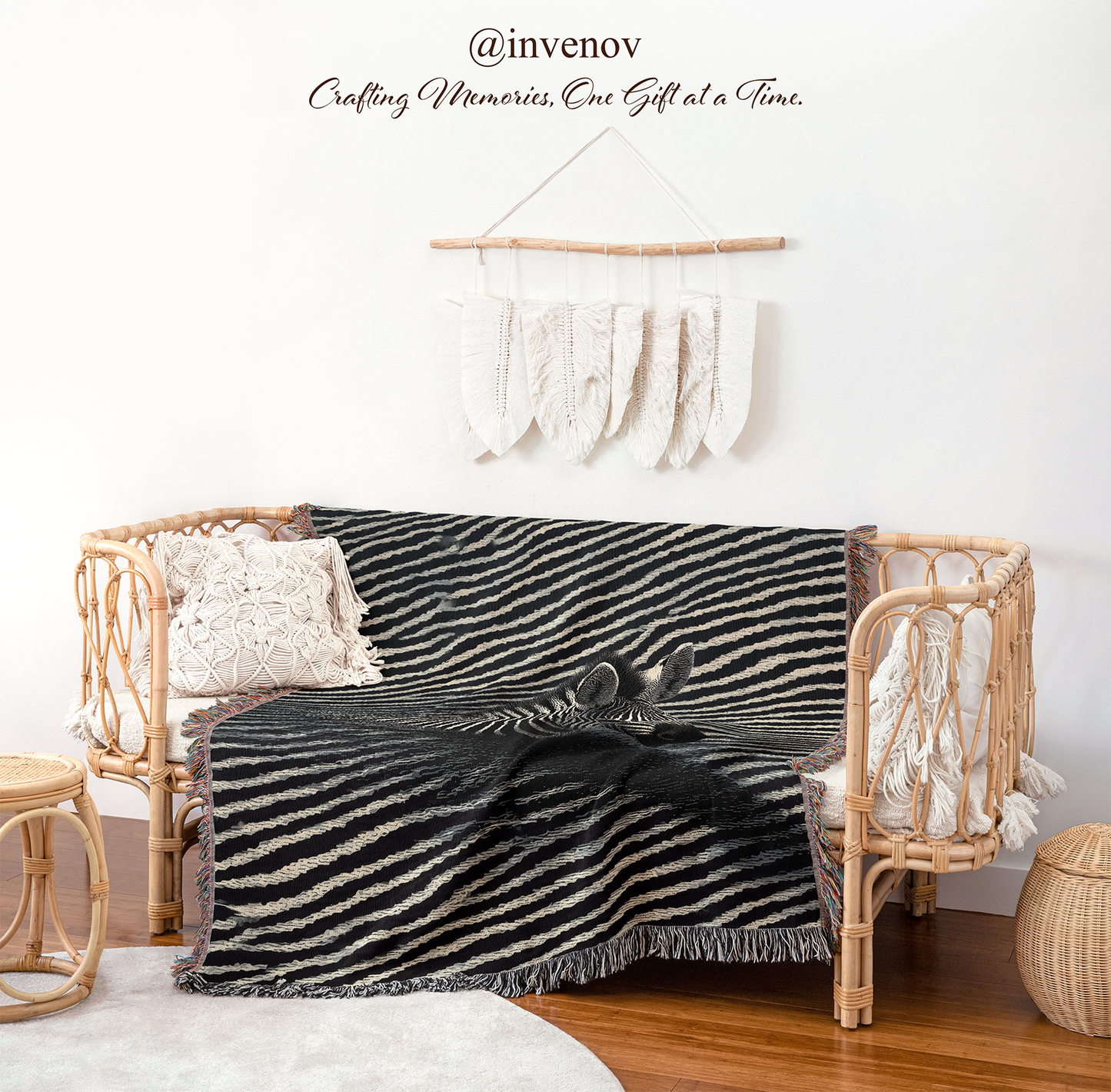 A black and beige zebra-print woven throw blanket with fringe is draped over a rattan daybed in a styled room setting. The scene includes a white wall with a macrame hanger, wooden flooring, a small woven stool, and decorative pillows.